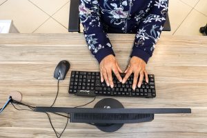 Foto tirada de cima, onde aparece só os braços de uma mulher, que está de casaco florido. As mãos dela estão sobre o teclado e ela está de frente para o computador, provavelmente trabalhando.