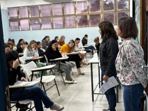 Sala cheia com os candidatos do concurso realizando a prova e duas examinadoras em pé, de frente para os candidatos.